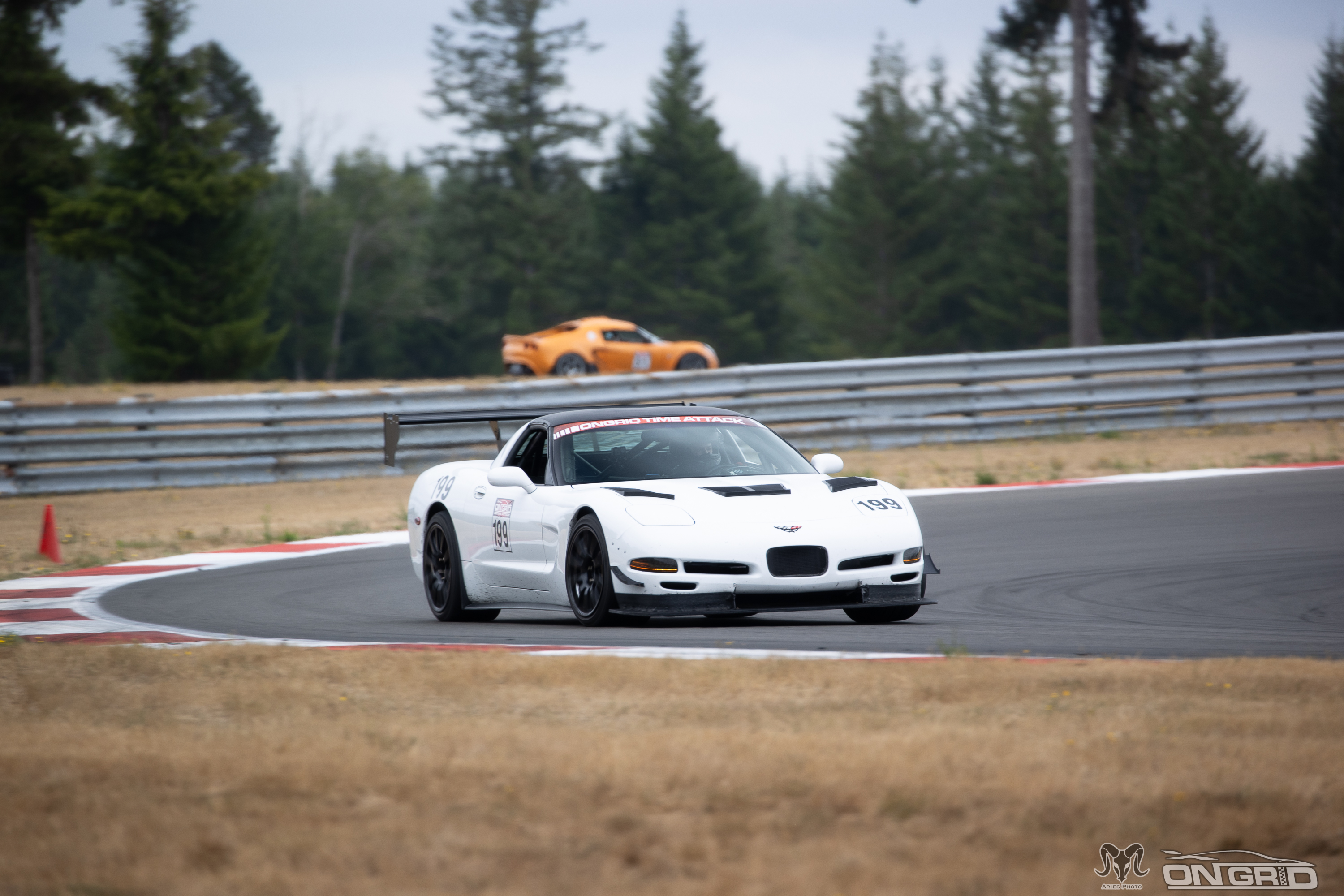 A white C5 Corvette mid corner with the OnGrid time attack banner on the car. In the distance is an orange car that isn't as fast as the white corvette. It's not their fault. They should have bought a Corvette.