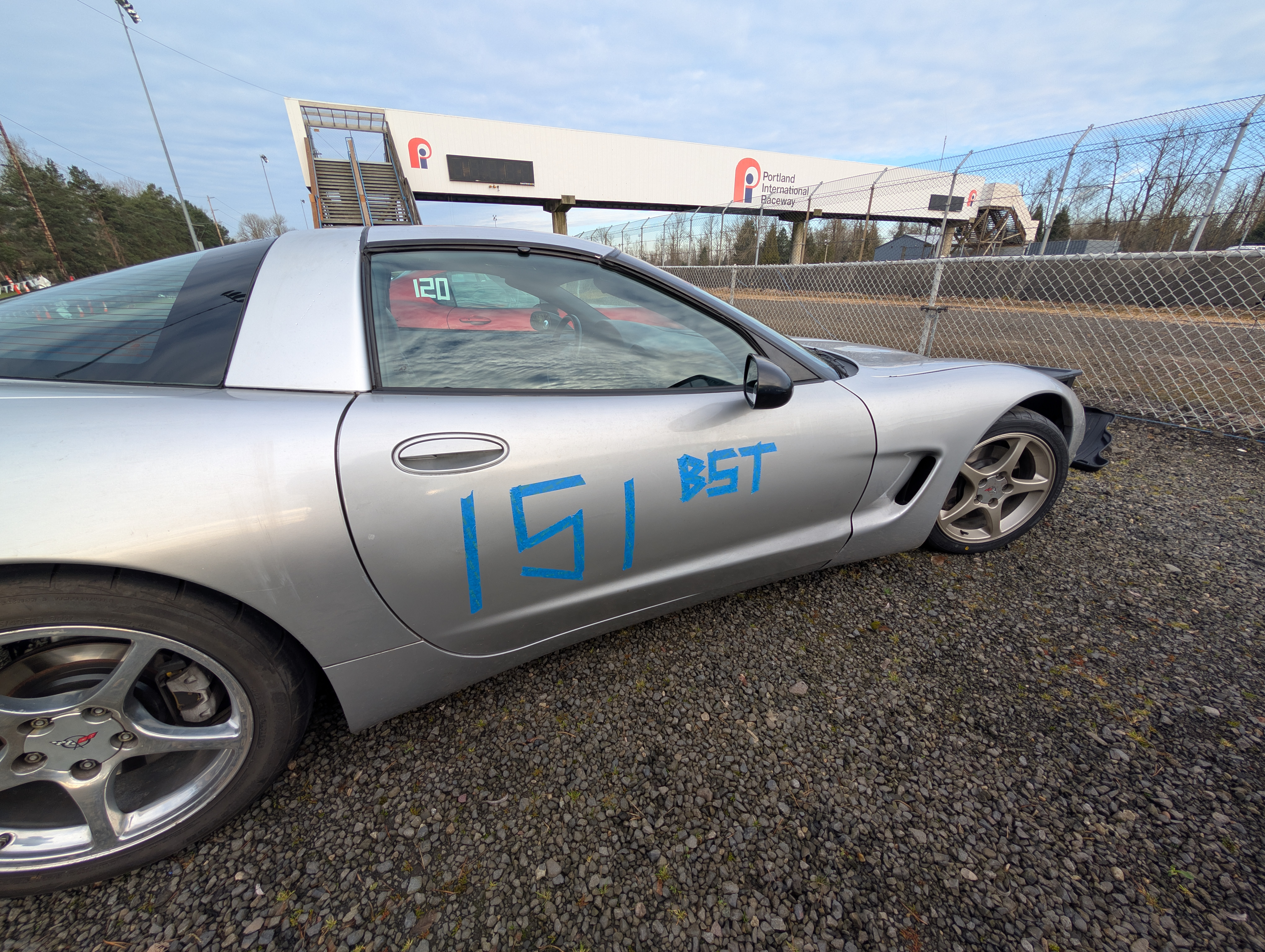 The Dead to Shred Corvette parking in the gravel by the front straight of PIR, with the pedestrian bridge sporting the PIR logo in the background. The car has "151 BST" written on the side with blue painters tape.